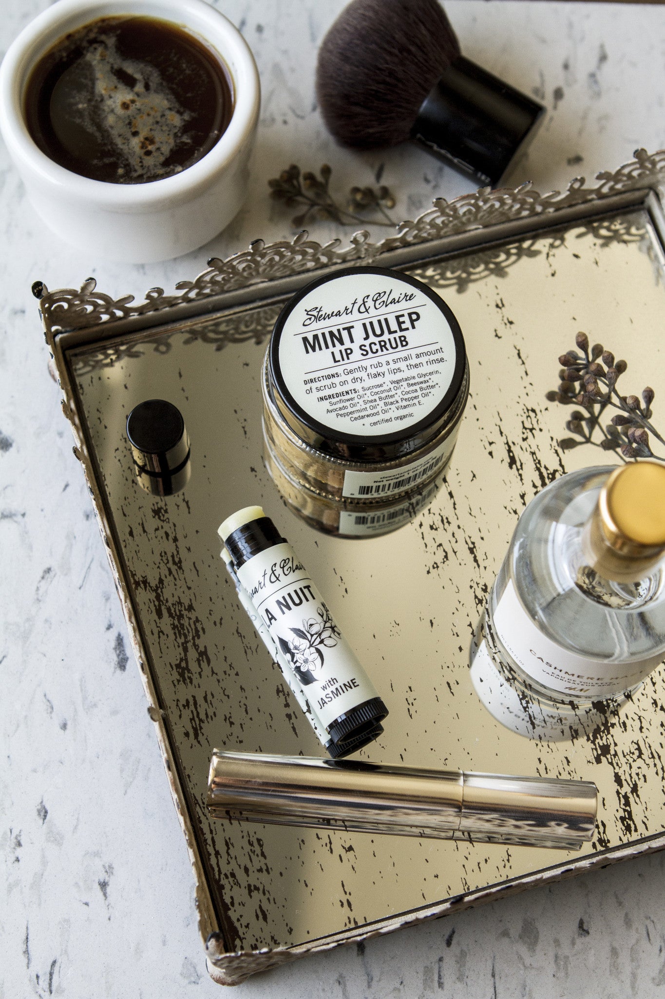 An overhead image of a jar of lip scrub and a lip balm on a bronze tray with mascara. In the background is a blush brush and a cup of coffee.