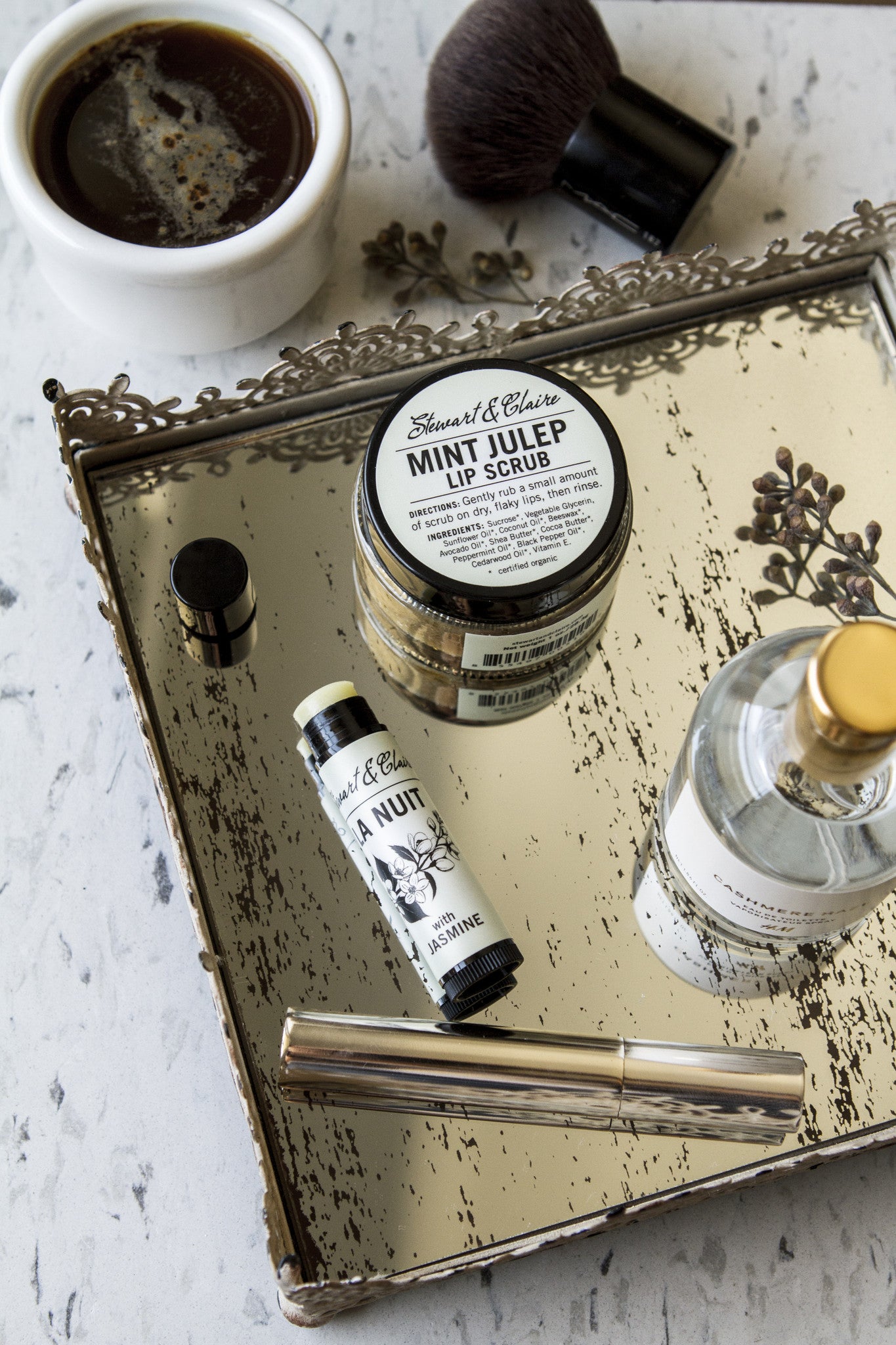 An overhead image of a jar of lip scrub and a lip balm on a bronze tray with mascara. In the background is a blush brush and a cup of coffee. 