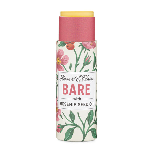 A .3 ounce paperboard tube of lip balm. The tube is off-white with illustrations of roses and rose hips. The tube is open at the top to reveal the balm, which is slightly pushed up. There is a white square on the tube with text that shows Stewart & Claire in script. It says Bare it large pink letters then with rosehip seed oil in black letters. It is on a white background.