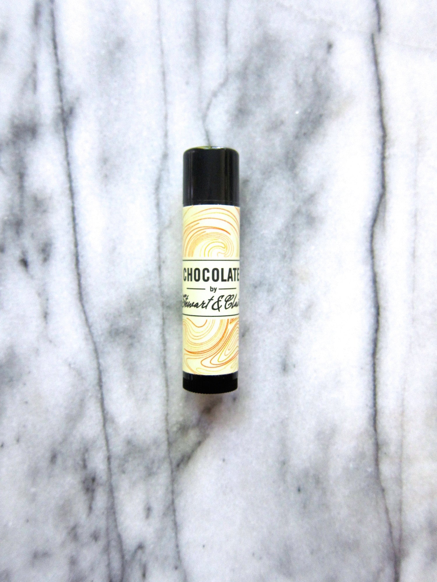 An image of a .15 ounce tube of lip balm. The tube is black plastic. The label is off-white with has a swirly pattern that has orange, brown, and slightly reddish lines. In the center, it says, "Chocolate by Stewart & Claire." The letters are black. The background is marble..