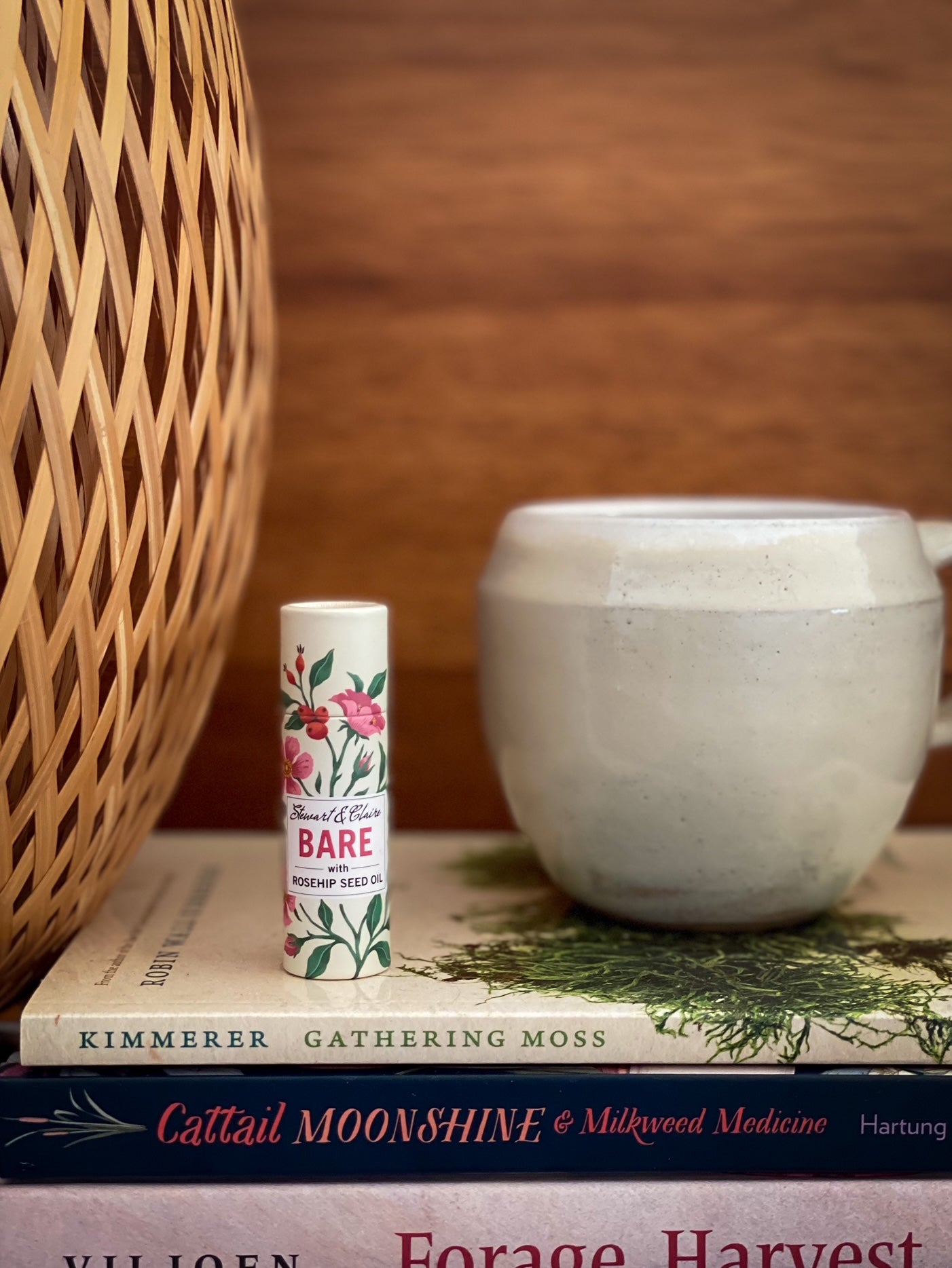 A .3 ounce paperboard tube of lip balm. The tube is off-white with illustrations of roses and rose hips. There is a white square on the tube with text that shows Stewart & Claire in script. It says Bare it large pink letters then with rosehip seed oil in black letters. It is sitting on a knitted cream-colored blanket. The balm is standing on a stack of books between a lamp with a honeycomb shade and a hand-made ceramic mug glazed in white.