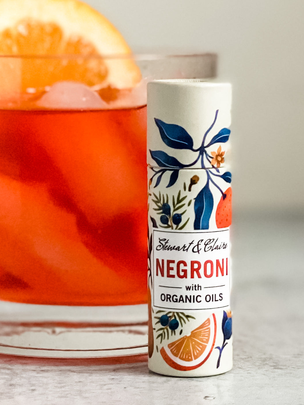 An image of a Negroni cocktail in a squat glass with an orange wheel in the background set on a gray surface. In front is an off white paperboard tube with botanical illustrations of oranges with blue leaves and juniper berries on juniper branches. Toward the center of the tube is a white box. At the top, it says Stewart & Claire in script. Underneath it says Negroni in red letters and then with organic oils in black letters. 