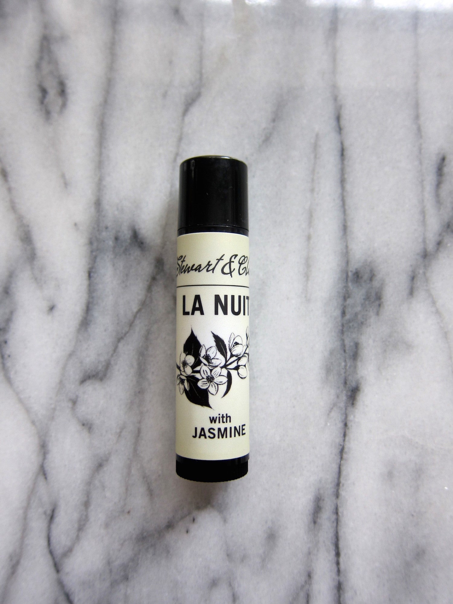 An image of a .15 ounce lip balm tube in black plastic. The tube has a an off-white label that says Stewart & Claire in black script. Then La Nuit in large black letters. Below that is a black and white illustration of jasmine and below it, the text says, with jasmine. It is on a marble background.