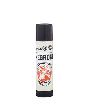 A .15 ounce black plastic tube of lip balm with an off-white label. The top of the label says Stewart & Claire in script. Black letters then say Negroni. Below is an illustration of a red cocktail that looks like a Negroni. The background is white.