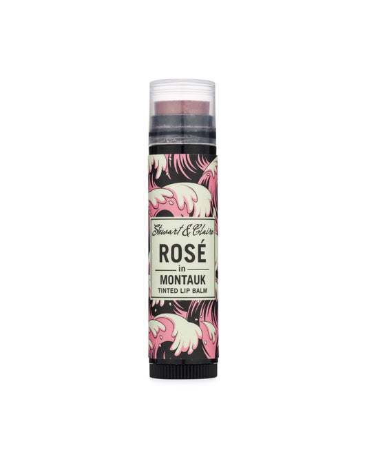 An image of a .15 ounce black plastic tube with a clear cap. Through the cap, you can see a lip balm that is a dusky pink. The tube has a label in pink, black, and white with a wave pattern. In the center of the balm is an off-white square that says Stewart & Claire in black script and then Rosé in Montauk, Tinted Lip Balm underneath.