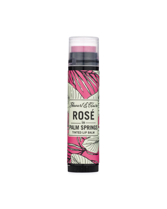 An image of a .15 ounce black plastic tube with a clear cap. Through the cap, you can see a lip balm that is a bright pink. The tube has a label in pink, black, and white with a leafy pattern. In the center of the balm is an off-white square that says Stewart & Claire in black script and then Rosé in Palm Springs, Tinted Lip Balm underneath.