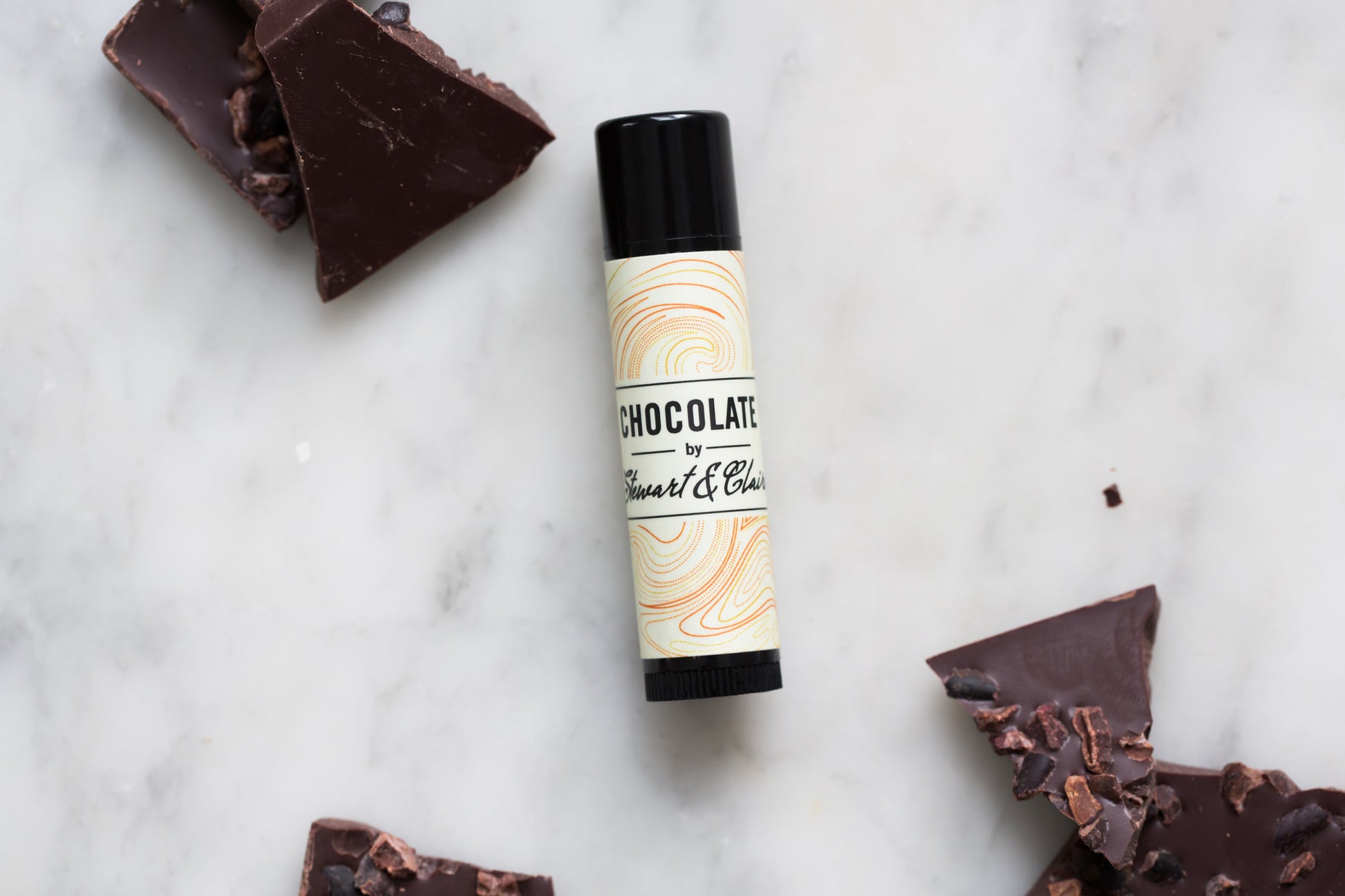An image of a .15 ounce tube of lip balm. The tube is black plastic. The label is off-white with has a swirly pattern that has orange, brown, and slightly reddish lines. In the center, it says, "Chocolate by Stewart & Claire." The letters are black. The background is marble and there are pieces of broken up dark chocolate bars with cocoa nibs in them also in the image.
