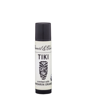An image of a .15-ounce black plastic lip balm tube with a white label. At the top, the label says Stewart & Claire in black script. underneath, it says Tiki. Below that is a tiki mask (note that this lip balm has been updated with an illustration showing a coconut.) It then says scented with Mandarin Orange.