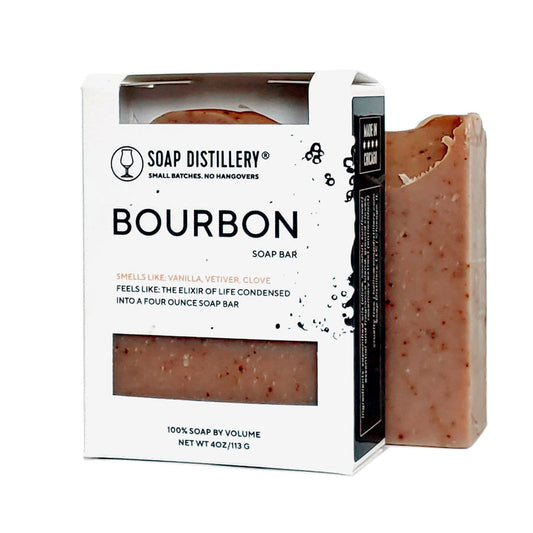 An image of a light brown soap with dark brown flecks outside a box alongside the same type of soap inside a box. The box is white in front with black sides. On the front, it says, Soap Distillery: Small Baches. No Hangovers. Bourbon Soap Bar. Smells like vanilla, vetiver, clove. Feels like the elixir of life condensed into a four ounce soap bar. 100% soap by volume. net wt 4 oz/113 g