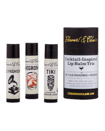 An image of the Stewart & Claire Cocktail-Inspired Lip Balm Trio. It shows three .15 ounce lip balms in black plastic tubes with off-white labels. From left-to-right, the lip balms are: Old Fashioned, Negroni, Tiki. To the right of those lip balms is a larger black paperboard tube. At the top, it says Stewart & Claire in gold foil script. At the bottom, it says stewartandclaire.com. In the center, there is a white label that says "Cocktail-Inspired Lip Balm Trio with a black illustration of a coupe.
