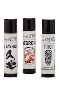 An image that shows three .15 ounce black plastic lip balm tubes, each with off-white labels. The labels each have Stewart & Claire in script at the top. From left-to-right, the balms are Old Fashioned, Negroni, Tiki.