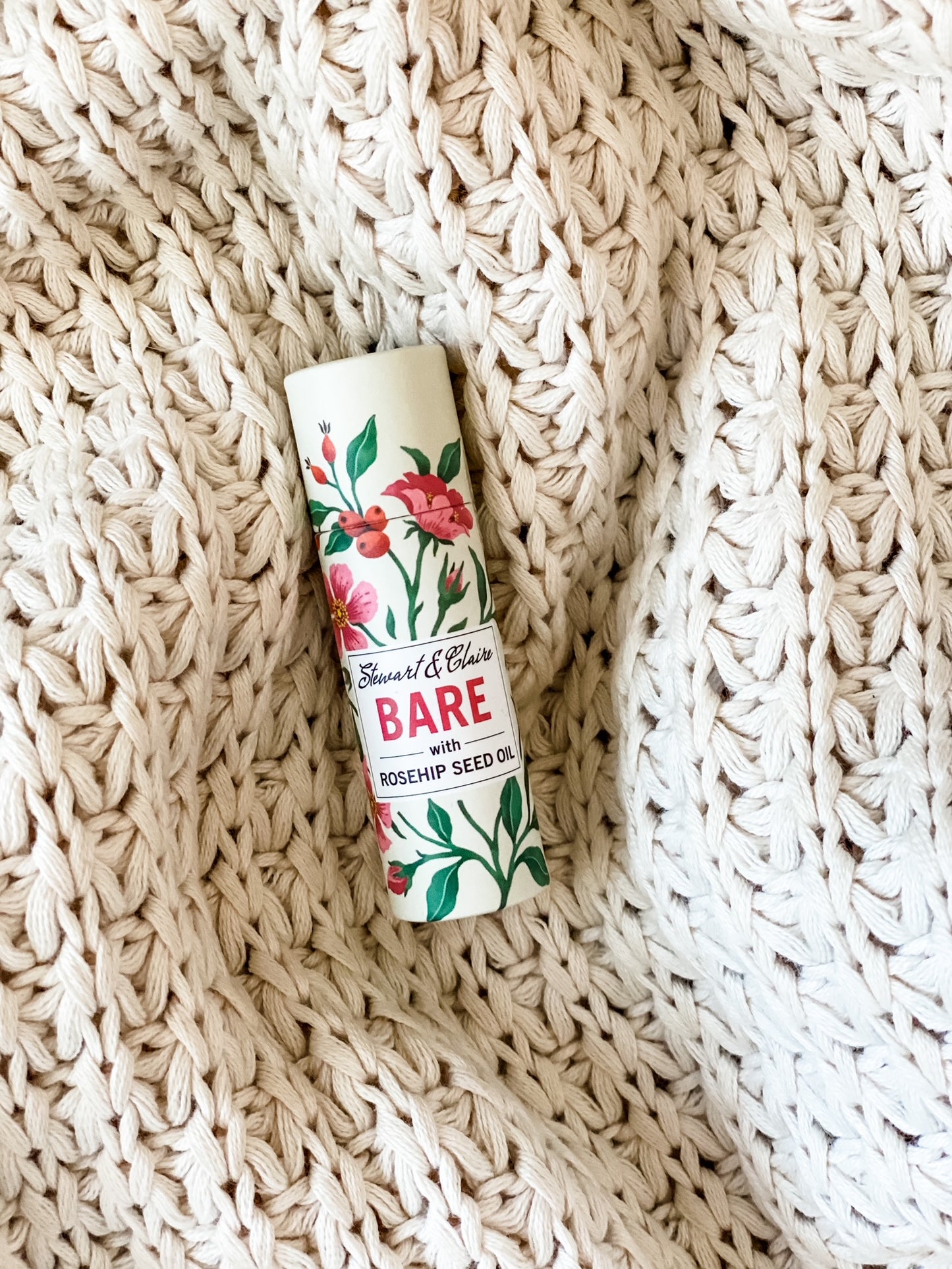A .3 ounce paperboard tube of lip balm. The tube is off-white with illustrations of roses and rose hips.  There is a white square on the tube with text that shows Stewart & Claire in script. It says Bare it large pink letters then with rosehip seed oil in black letters. It is sitting on a knitted cream-colored blanket.