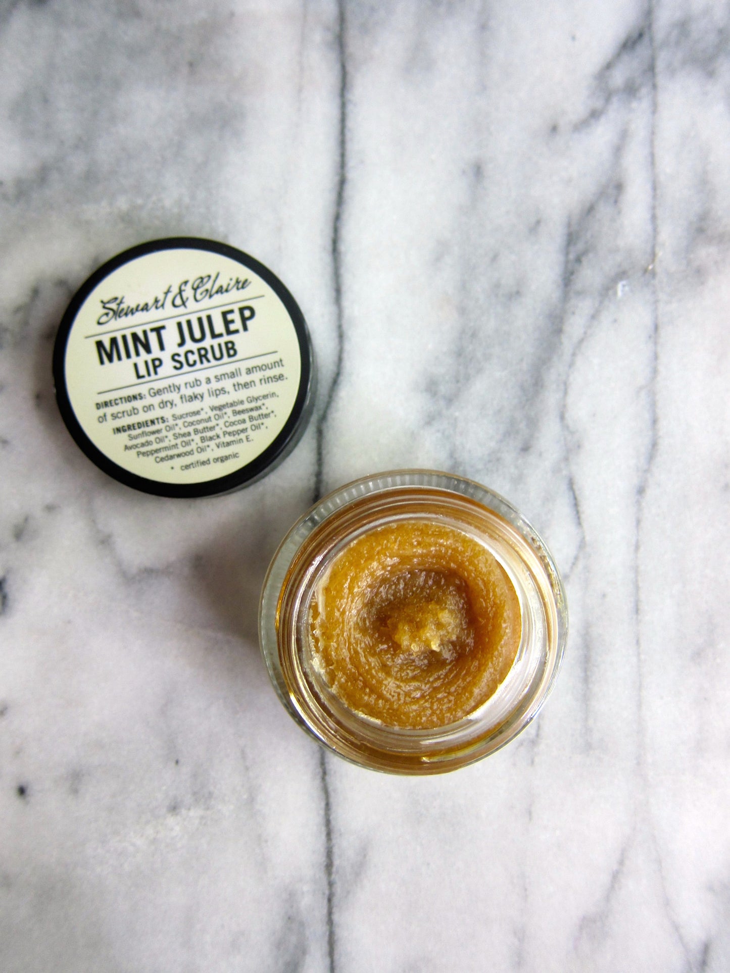 An image shot from overhead of a small squat glass jar with a black lid. The lid is off the jar revealing the brown lip scrub within. The off-white label on top of the jar  Stewart & Claire Mint Julep Lip Scrub. The text below shows instructions for use and the ingredients. 