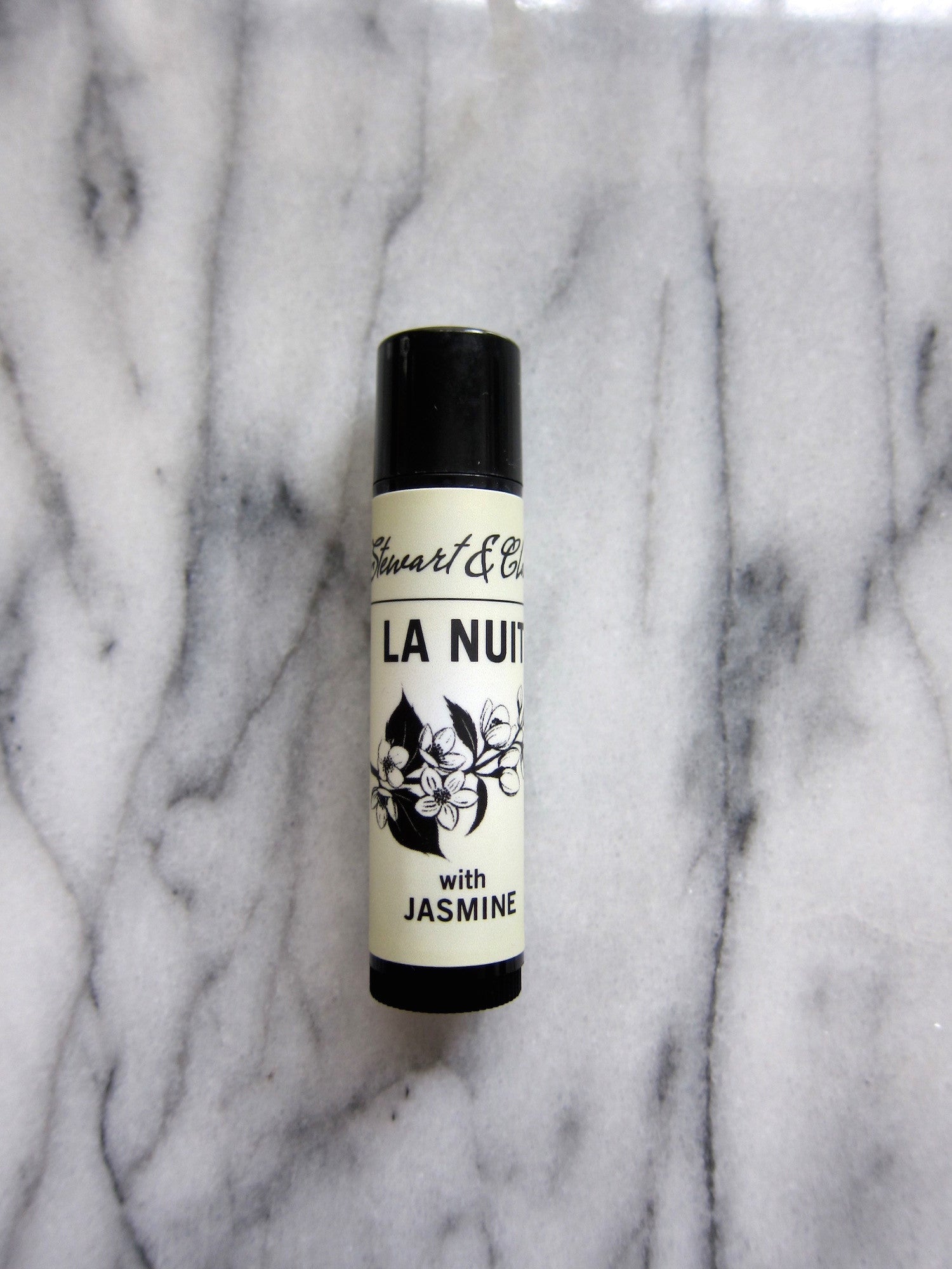 An image of a .15 ounce lip balm tube in black plastic. The tube has a an off-white label that says Stewart & Claire in black script. Then La Nuit in large black letters. Below that is a black and white illustration of jasmine and below it, the text says, with jasmine. It is on a marble background. 