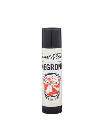A .15 ounce black plastic tube of lip balm with an off-white label. The top of the label says Stewart & Claire in script. Black letters then say Negroni. Below is an illustration of a red cocktail that looks like a Negroni. The background is white. 