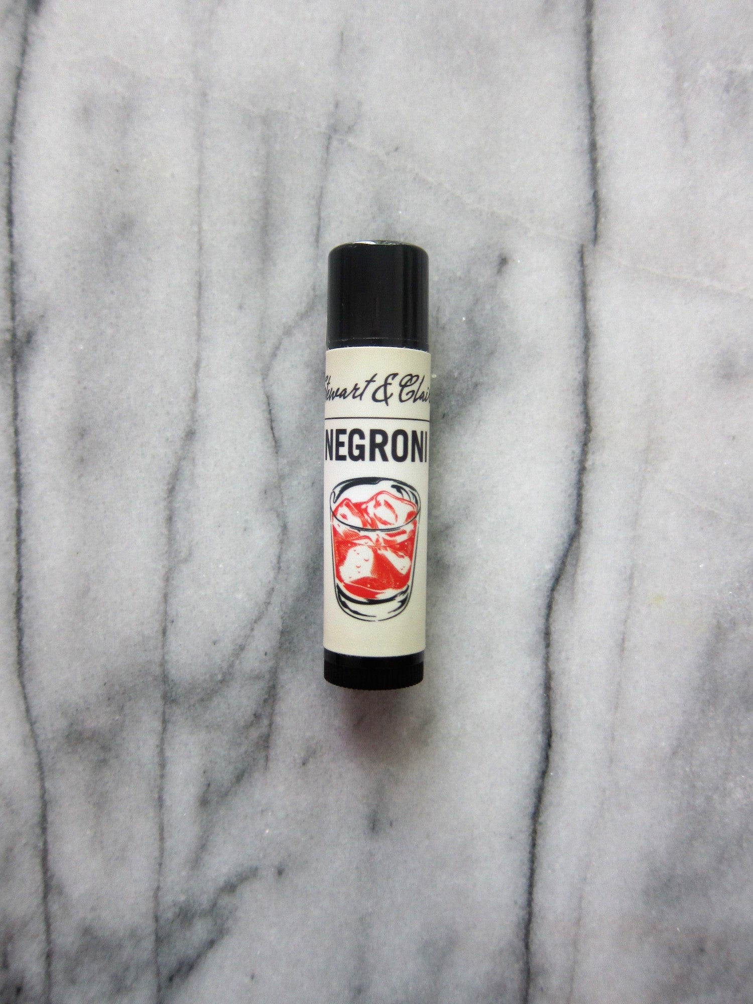 A .15 ounce black plastic tube of lip balm with an off-white label. The top of the label says Stewart & Claire in script. Black letters then say Negroni. Below is an illustration of a red cocktail that looks like a Negroni. The background is marble. 