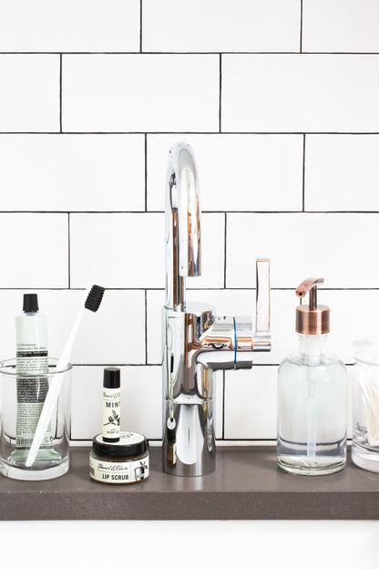 An image of a bathroom sink setting with a gooseneck chrome faucet. To the left of the faucet is a glass with toothpaste and a white toothbrush. To the right of that is a jar of Stewart & Claire lip scrub topped with Stewart & Claire Mint lip balm (now discontinued.) To the right of the faucet is a glass container of handsoap with a copper pump. The background is white subway tile with black grout and the sink rim is dark gray.
