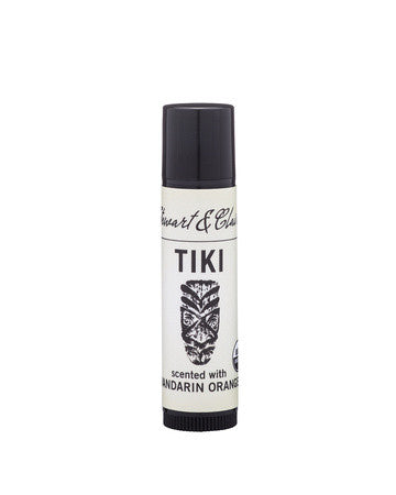 An image of a .15-ounce black plastic lip balm tube with a white label. At the top, the label says Stewart & Claire in black script. underneath, it says Tiki. Below that is a tiki mask (note that this lip balm has been updated with an illustration showing a coconut.) It then says scented with Mandarin Orange. 